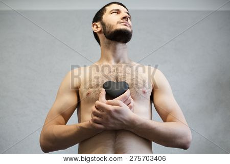 Naked Young, Bearded Man, Looking Up And Holding Black Heart In Hands Near Torso.