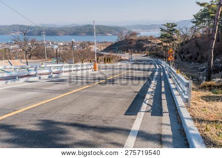Landscape Of Country Road Leading To Reservoir On Hazy Overcast Day With Mountains, City Buildings A
