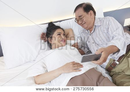 Happy Senior Vietnamse Couple Lying On Hotel Room And Watching Videos On Smartphone