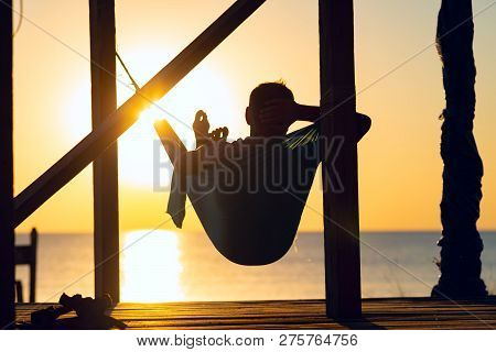 Man Relaxes In A Hammock On Abandoned Bungalow Veranda, Admiring The Turquoise Sea During Sun Set. A