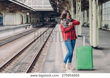 Photo Of Attractive Young Woman Waits Train At Railway Station, Dressed In Knitted Sweater And Jeans