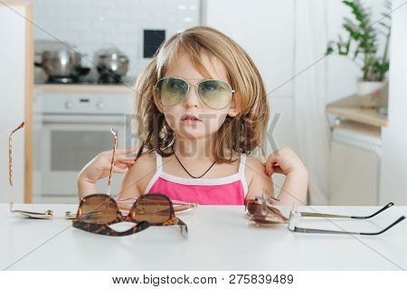 Portrait Of Cute Dark Blonde Little Girl With Short Hair Trying To Wear Blue Aviator Sunglasses. She