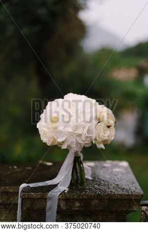 Minimalistic Bridal Bouquet Of White Roses And Hortense With White Ribbons On The Wet Stone Railing