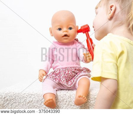 Little Girl Inspects The Ears Of Her Doll On A White Background. The Concept Of Pediatric Otolaryngo