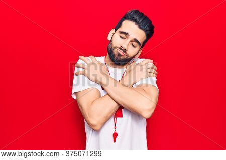 Young handsome man with beard wearing lifeguard t-shirt and whistle hugging oneself happy and positive, smiling confident. self love and self care 