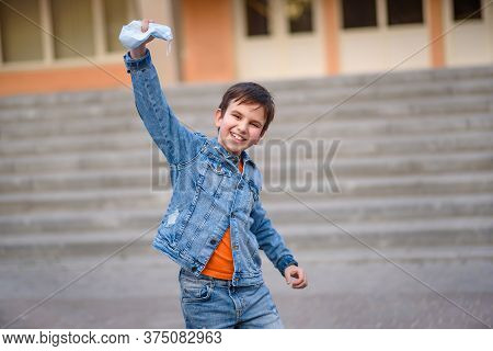 Happy Smiling Schoolboy  Takes Off His Protective Maskin Front Of School Stairs. End Of Pandemic Cor