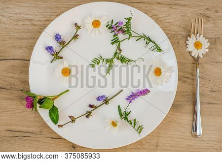 Flowers And Green Plant In A White Plate And Fork On A Wooden Table, Vegetarian Food Concept.