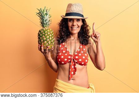 Middle age beautiful woman wearing bikini and summer hat holding pineappel smiling with an idea or question pointing finger with happy face, number one 