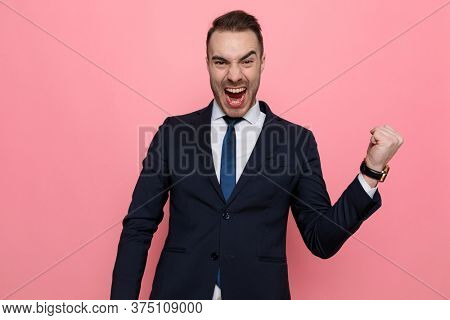 excited young businessman in suit holding fist in the air and celebrating victory, screaming and standing on pink background