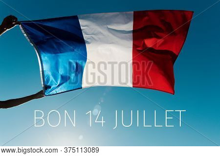man holding a French flag waving on the wind and the text bon 14 juillet, happy 14 july, the national day of France written in French, against the blue sky, in backlit