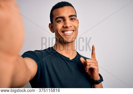 Young handsome african american man wearing casual t-shirt making selfie by camera surprised with an idea or question pointing finger with happy face, number one