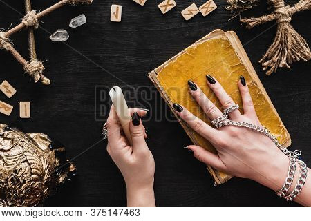Top View Of Woman Holding Candle Near Aged Book, Voodoo Doll, Runes, Crystals And Skull On Black
