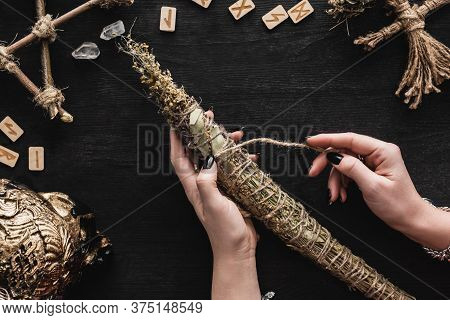 Top View Of Woman Holding Dried Herbs Near Runes, Voodoo Doll, Runes, Skull And Crystals On Black