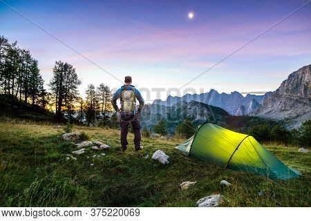 Hiker With Backpack Standing On Mountain And Tent Glowing Under A Moon Night Sky At Sunset Or Sunris