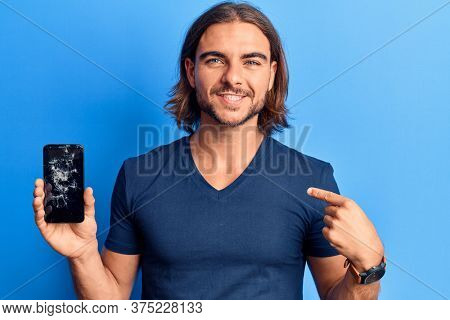 Young handsome man holding broken smartphone showing cracked screen pointing finger to one self smiling happy and proud 