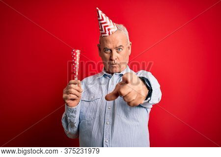 Middle age man wearing funny hat using trumpet celebrating birthday over red background pointing with finger to the camera and to you, hand sign, positive and confident gesture from the front