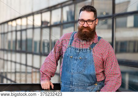 Portrait Of Brutal Bearded Stylish Craftsman Wearing Blue Overalls, Checked Shirt In Vintage Style O