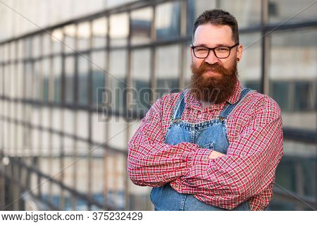 Portrait Of Brutal Bearded Stylish Craftsman Wearing Blue Overalls, Checked Shirt In Vintage Style O