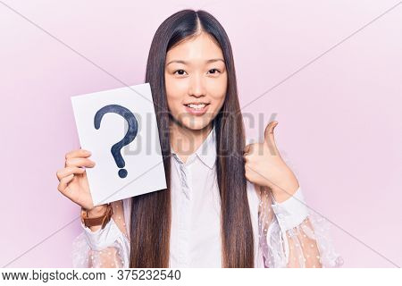 Young beautiful chinese woman holding question mark smiling happy and positive, thumb up doing excellent and approval sign 