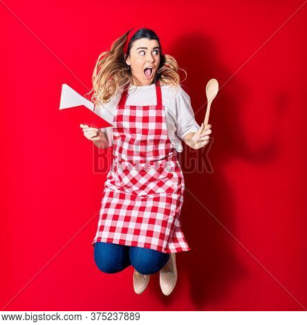 Young beautiful blonde cook woman wearing apron smiling happy. Jumping with smile on face holding recipe book and spoon over isolated red background