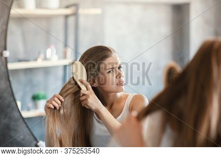 Frustrated Young Woman Trying To Brush Her Tangled Hair In Front Of Mirror At Bathroom