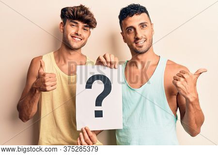 Young gay couple holding question mark smiling happy and positive, thumb up doing excellent and approval sign 