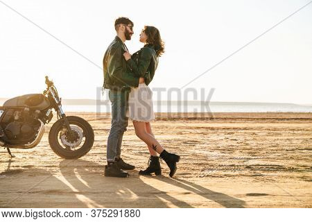 Beautiful young cople wearing jackets spending time at the sunny beach, standing near a motorbike