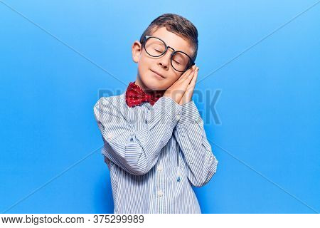 Cute blond kid wearing nerd bow tie and glasses sleeping tired dreaming and posing with hands together while smiling with closed eyes. 
