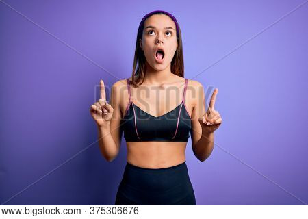 Young beautiful sporty girl doing sport wearing sportswear over isolated purple background amazed and surprised looking up and pointing with fingers and raised arms.