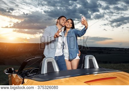 Preety Young Couple Going On A Long Drive In A Convertible Car. Couple Use Smartphone For Selfie.