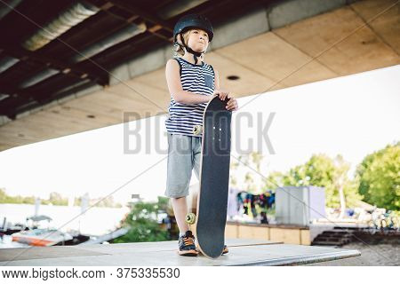 Young Boy Stands At Edge Of Half Pipe Ramp At Skateboard Park. Trendy Skateboarder Enjoying Free Tim