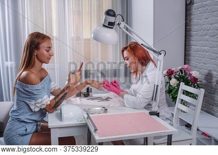 The Girl Sits In The Office On A Manicure Procedure. Young Woman Does Manicure In Salon