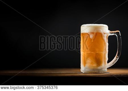 Cold Mug With Beer, With Overflowing Foam, On Wooden Table And Dark Background