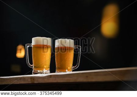 Two Cold Mugs With Beer, With Overflowing Foam, On Wooden Table And Dark Background, Space For Writi