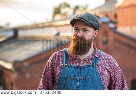 Portrait Of Brutal Bearded Stylish Craftsman Wearing Blue Overalls, Checked Shirt And Cap In Vintage