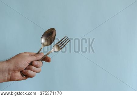 Hand Of An Adult Male Holds A Metal Spoon And Fork. Stainless Steel Spoon And Fork In The Left Hand.