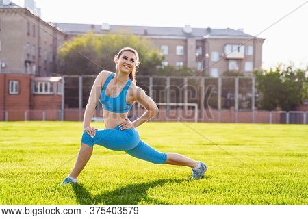 Attractive, Athletic Girl In A Blue Suit Stretches In The Stadium From Green Grass. Enthusiastically