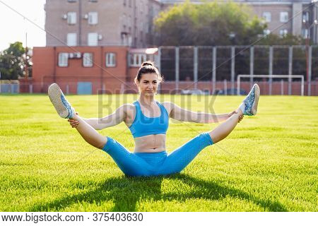 Beautiful, Athletic Caucasian Girl Sitting On A Twine In A Green Grassy Sports Stadium. Making Sport