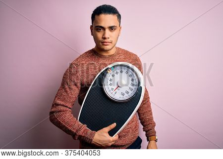 Young brazilian man doing diet to lose weigth holding scale over isolated pink background with a confident expression on smart face thinking serious