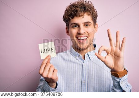 Young blond romantic man with curly hair holdiing paper with love message for valentines day doing ok sign with fingers, excellent symbol
