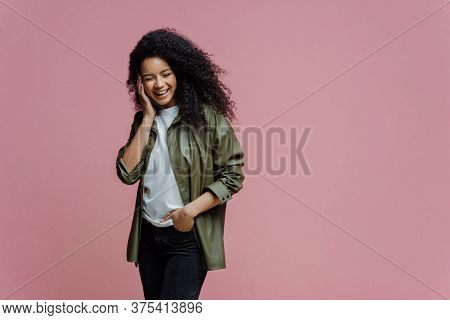 Horizontal Shot Of Happy African American Woman Keeps Hand In Pocket, Laughs As Hears Funny Anecdote