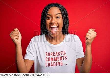 Young african american woman wearing sarcasm coments text on t-shirt over red background screaming proud and celebrating victory and success very excited, cheering emotion