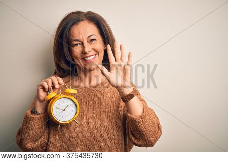 Middle age brunette woman holding clasic alarm clock over isolated background Waiving saying hello happy and smiling, friendly welcome gesture