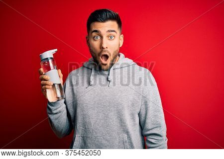 Young handsome sportsman drinking bottle of water to refeshment over red background scared in shock with a surprise face, afraid and excited with fear expression