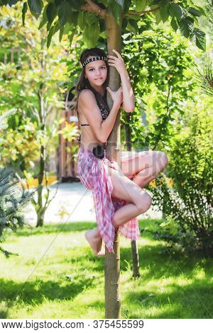 Teen Girl In A Swimsuit And Poreo Climbed A Tree. Portrait Of A Glamorous Beauty In A Summer Park.