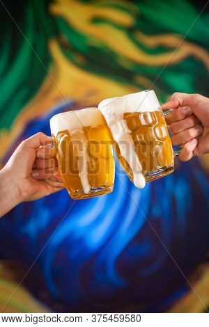 Two People Toasting With Mugs Full Of Draft Beer With Stylized Brazilian Flag Background