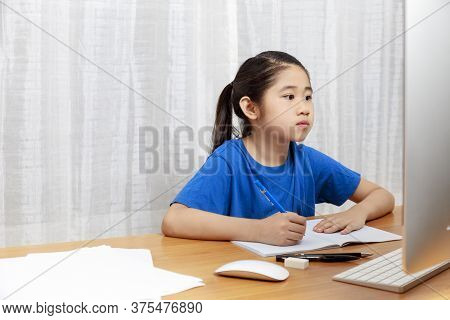 Asian Little Girl Is Studying Online Via The Internet  Sitting And Writing In  Living Room At Home. 