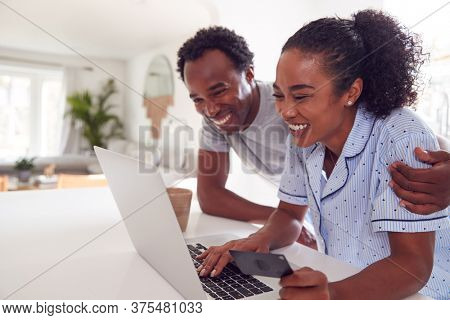 Couple Wearing Pyjamas In Kitchen Making Online Credit Card Purchase On Laptop