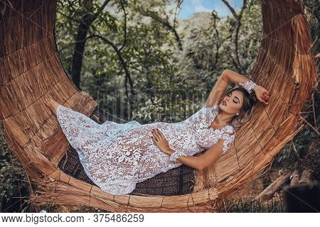 Beautiful Bride In Her Wedding Dress Posing For Photos On A Wooden Swing In The Tree.