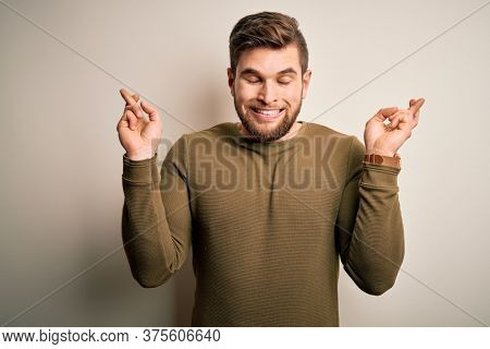 Young blond man with beard and blue eyes wearing green sweater over white background gesturing finger crossed smiling with hope and eyes closed. Luck and superstitious concept.
